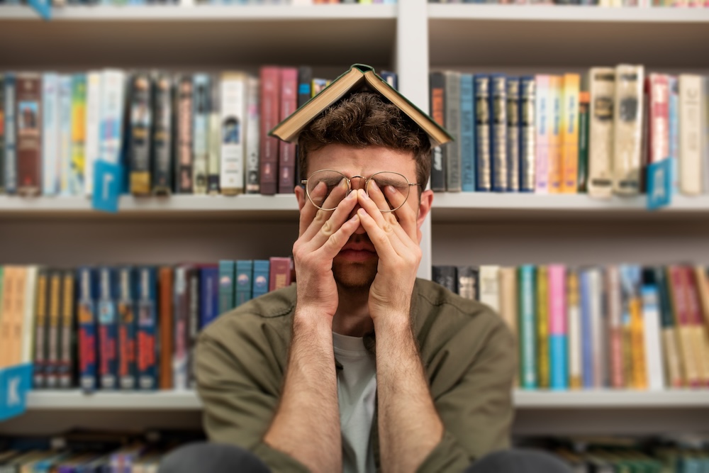 University student has difficulty to study, rubbing his eyes with a book on his head